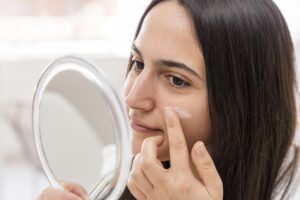 Close-up of a woman applying moisturizer to her cheek while looking in a mirror, illustrating skin barrier repair and hydration.